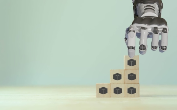 Robotic hand placing a wooden block with a cube icon on top of a pyramid of similar blocks against a pale blue-green background